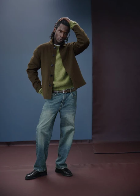 Full-length studio shot of an adult male model with dreadlocks in a green sweater, brown jacket, blue jeans, and black shoes.