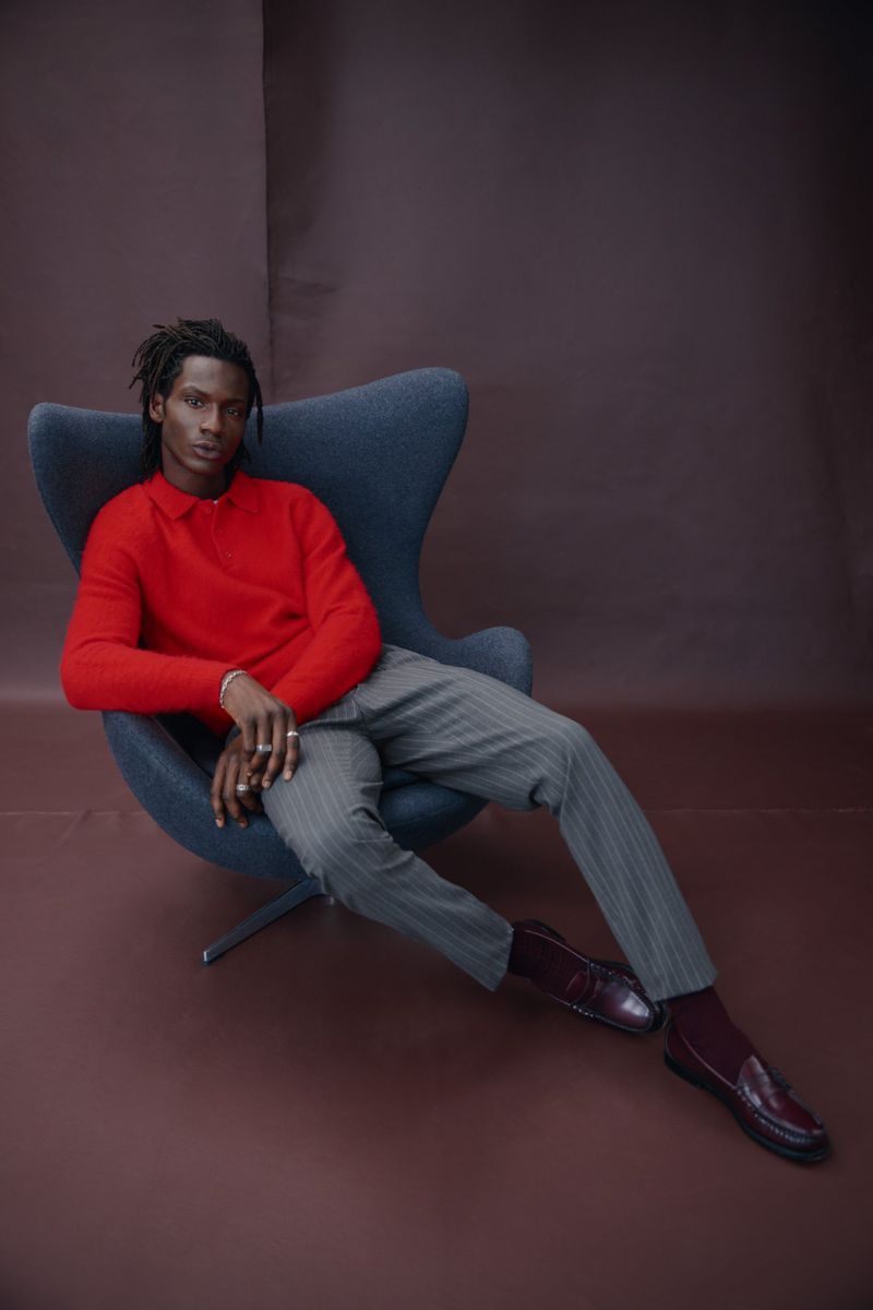 An adult male model with dreadlocks in a red sweater and grey trousers sits in a blue chair against a brown backdrop in a studio.