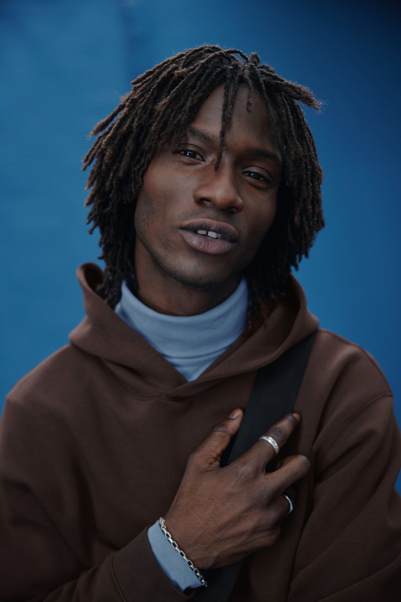 Portrait of a young adult Black man with dreadlocks, wearing a brown hoodie and grey turtleneck against a blue background in studio lighting.