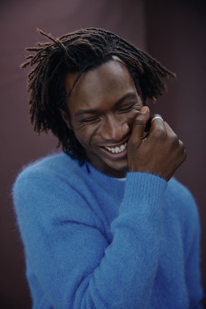 Portrait of an African-American man with dreadlocks smiling while wearing a blue sweater in front of a brown backdrop.