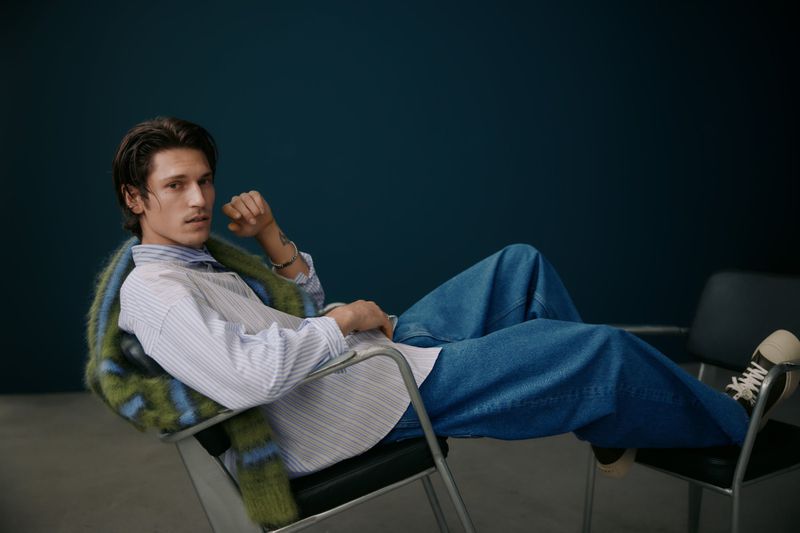 A young adult man with dark hair poses in blue jeans and a striped shirt, seated on a chair against a dark teal background.