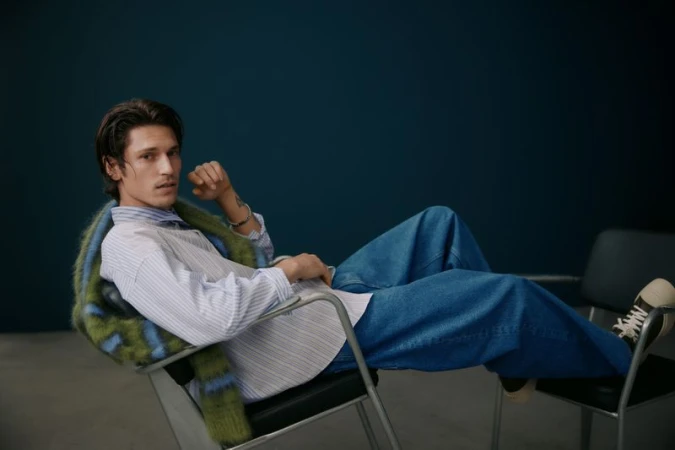 A young adult man with dark hair poses in blue jeans and a striped shirt, seated on a chair against a dark teal background.