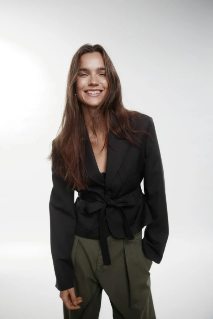 Smiling adult woman poses in a black blazer with a tie waist and green pants against a bright white backdrop in a studio setting.