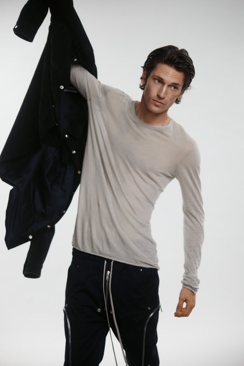 Adult male fashion model in light gray long sleeve shirt and black pants holds black jacket in a light gray studio setting.