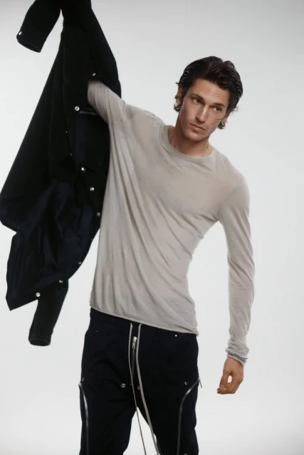 Adult male fashion model in light gray long sleeve shirt and black pants holds black jacket in a light gray studio setting.