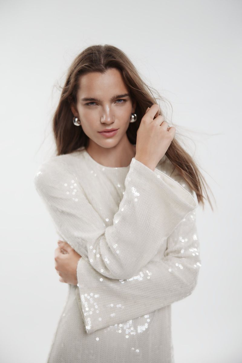 Model Laura Helene Christensen wearing a white sequined long-sleeve dress and silver ball earrings against a light studio backdrop.
