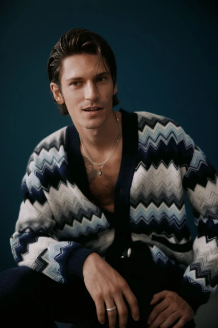Portrait of a young adult man in a patterned cardigan. He is wearing a silver chain and ring, and is against a teal background.