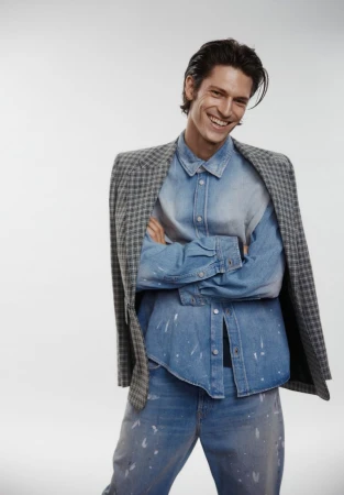 Smiling adult male in denim shirt, jeans, and grey patterned blazer, arms crossed in a studio shot against a white backdrop.