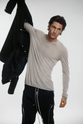 Male model in a light grey long-sleeve shirt and black zip trousers holding a dark jacket aloft against a white studio backdrop.