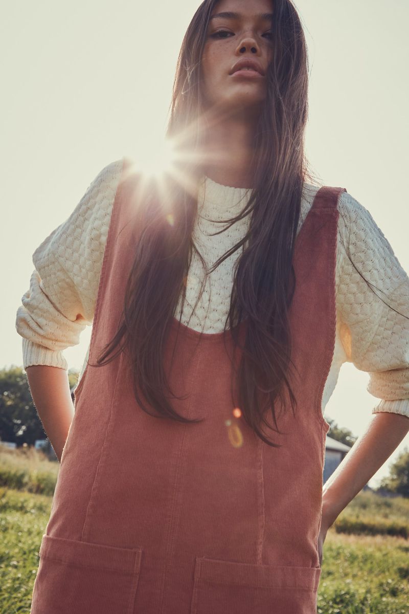 Young woman with long brown hair is the primary subject in a low-angle shot in a rural setting wearing a cream-colored sweater and a red dress.