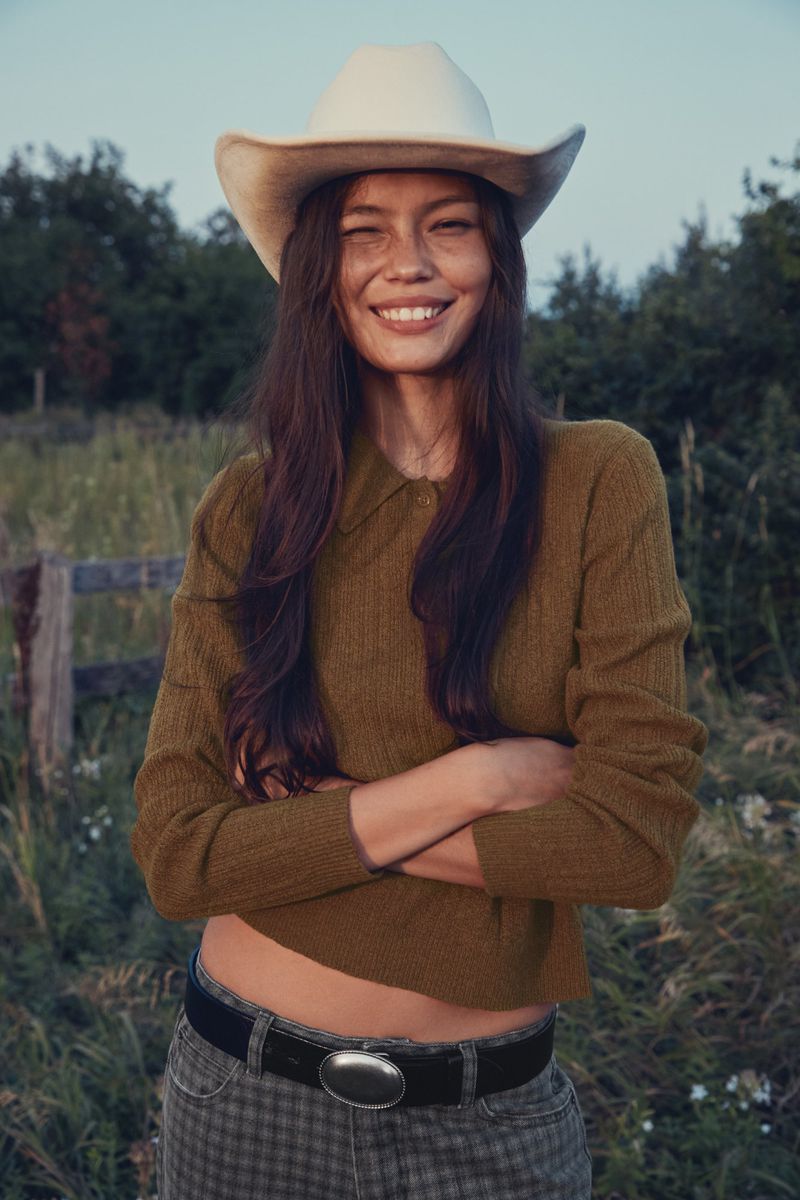 A young woman in a cowboy hat, brown shirt, and jeans stands smiling with her arms crossed in a grassy field, trees in the background.