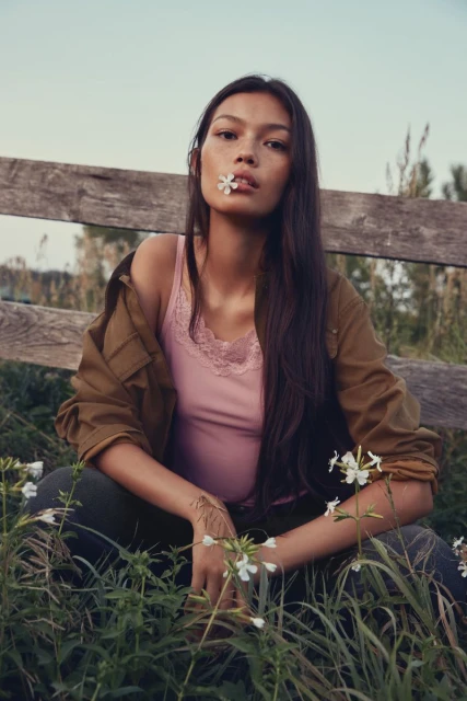 Young woman sits in field of grass and wildflowers wearing brown jacket and pink top with white flower in her mouth.