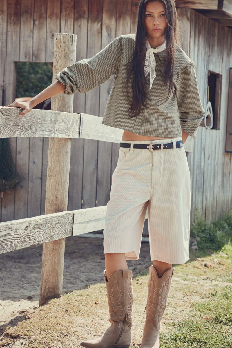 A woman poses near a wooden fence wearing a plaid shirt, white shorts, cowboy boots, and a white scarf against a rural backdrop.