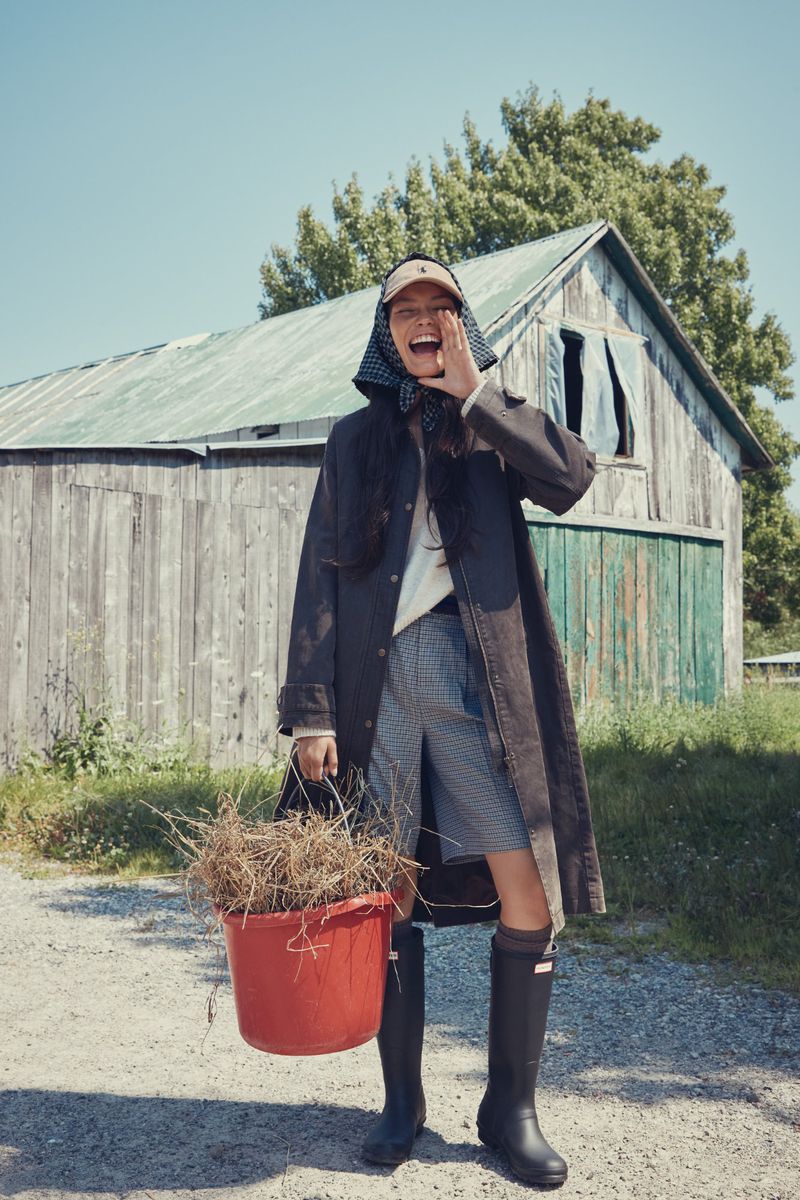 Woman in coat, headscarf, and boots laughs while holding a bucket of dry foliage in front of a weathered barn and clear sky.