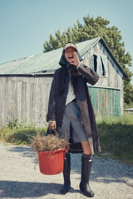 Woman in coat, headscarf, and boots laughs while holding a bucket of dry foliage in front of a weathered barn and clear sky.