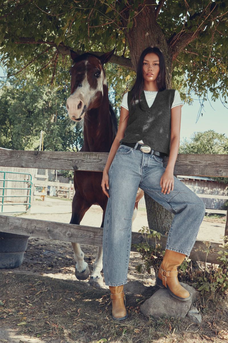 Heather Diamond Strongarm leaning against a wooden fence beside a brown and white horse wearing a knit vest and jeans.