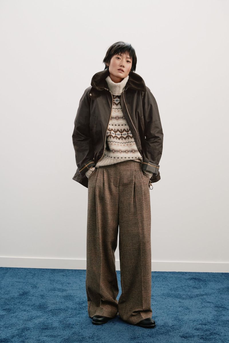Woman posing in a studio wearing a brown jacket, a patterned sweater, and pleated brown plaid pants against a white wall and blue floor.