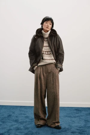 Woman posing in a studio wearing a brown jacket, a patterned sweater, and pleated brown plaid pants against a white wall and blue floor.