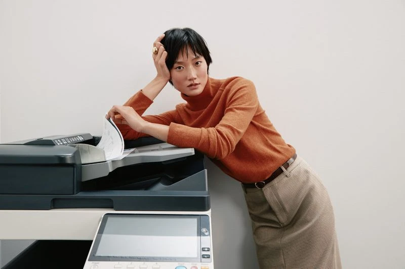 Woman leans on a black multifunction printer wearing an orange turtleneck sweater and checkered midi skirt in an indoor office environment.
