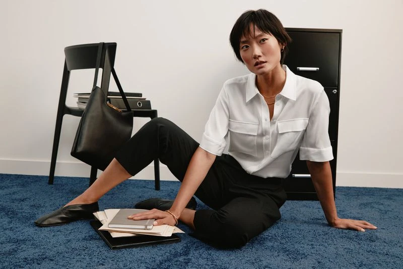 Adult woman in a white shirt and black pants sits on a blue carpet with a chair, filing cabinet, papers, and a black tote bag.