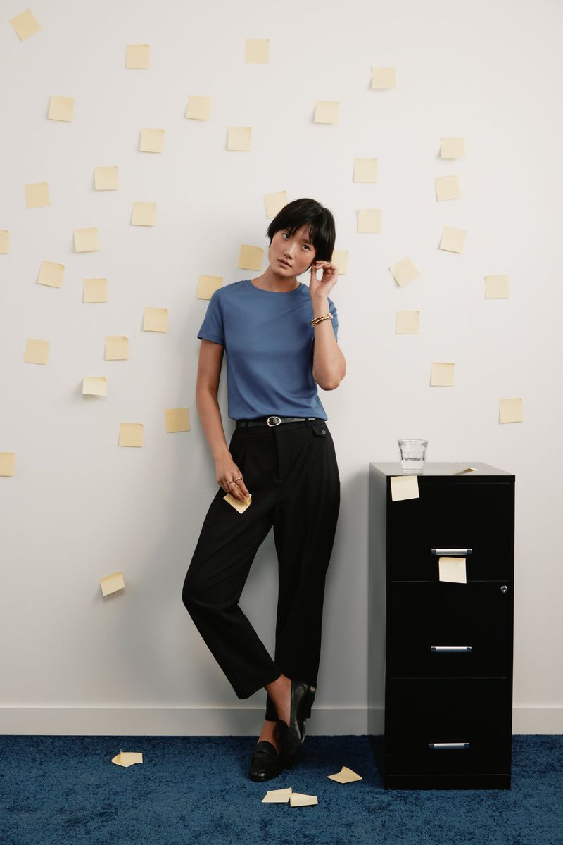 Woman in a blue top and dark pants stands in front of a white wall covered in yellow sticky notes, next to a black filing cabinet.