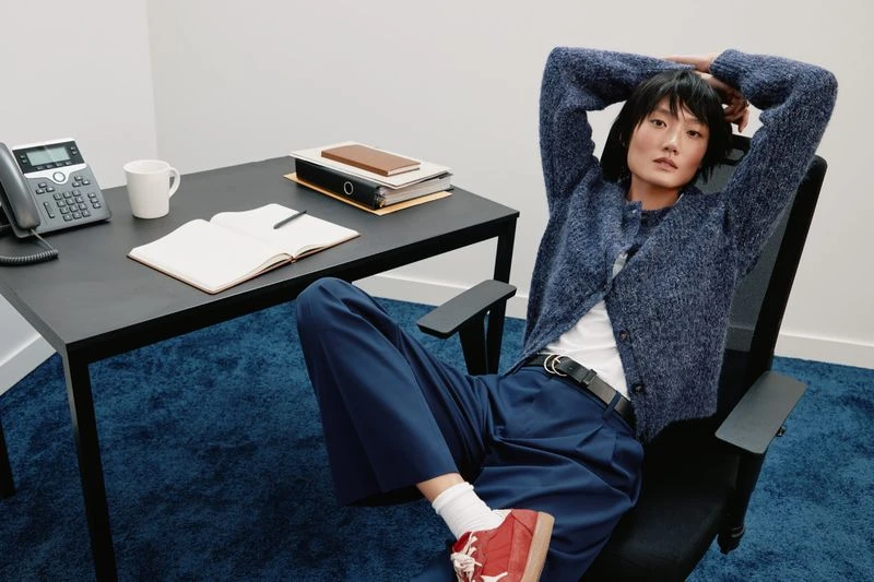 A young adult woman sits at an office desk in a blue sweater and pants. Desk items include a phone, mug, notebook, and files.