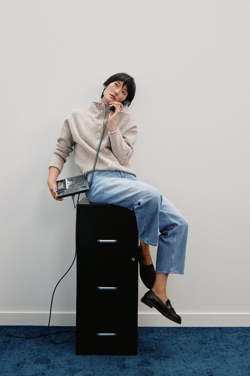 Woman in casual fashion sits atop a black file cabinet holding a vintage telephone in a minimalist white indoor setting.