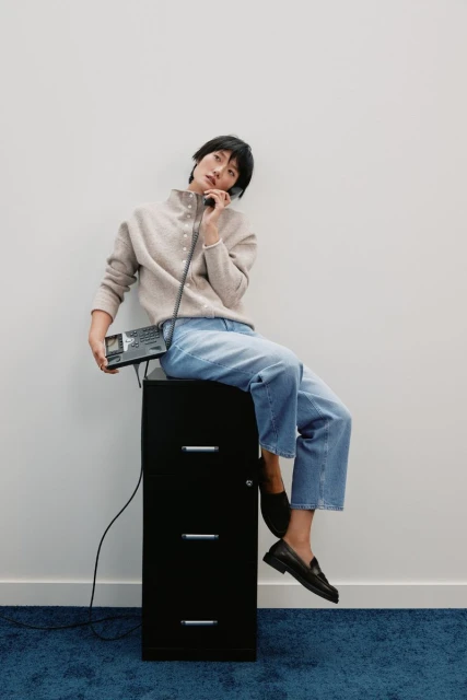 Woman in casual fashion sits atop a black file cabinet holding a vintage telephone in a minimalist white indoor setting.