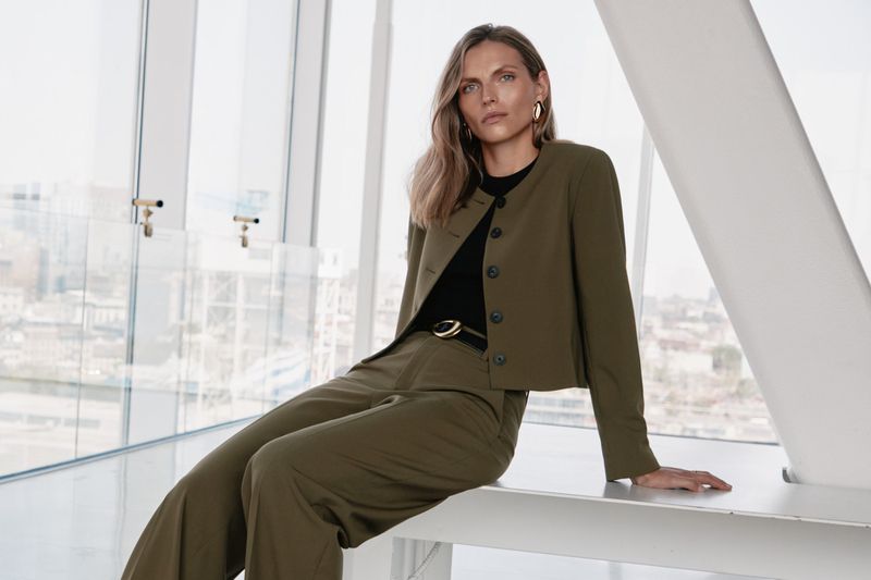 Blonde adult female model in olive green suit sitting indoors against a bright window with city view in fashion editorial shot.