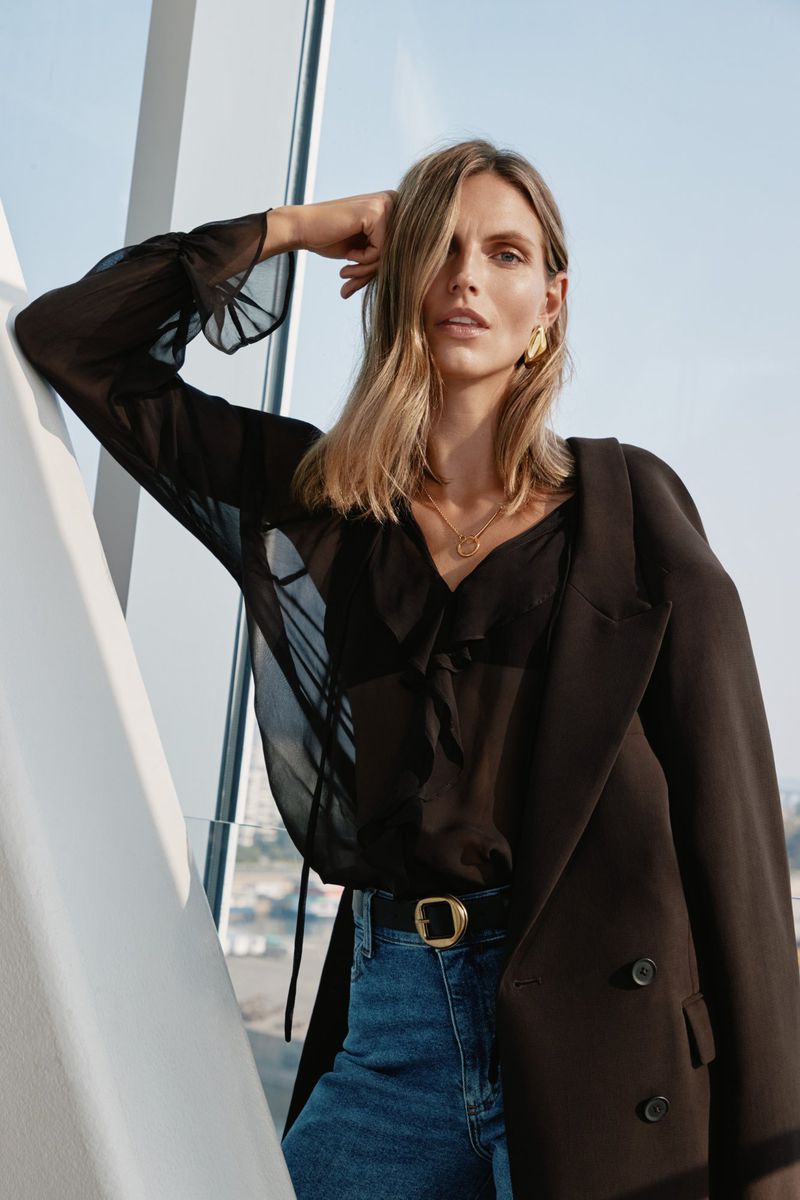 Elegant woman in a black blouse and blazer poses near a window, showcasing fashion and style with a confident gaze and chic attire.
