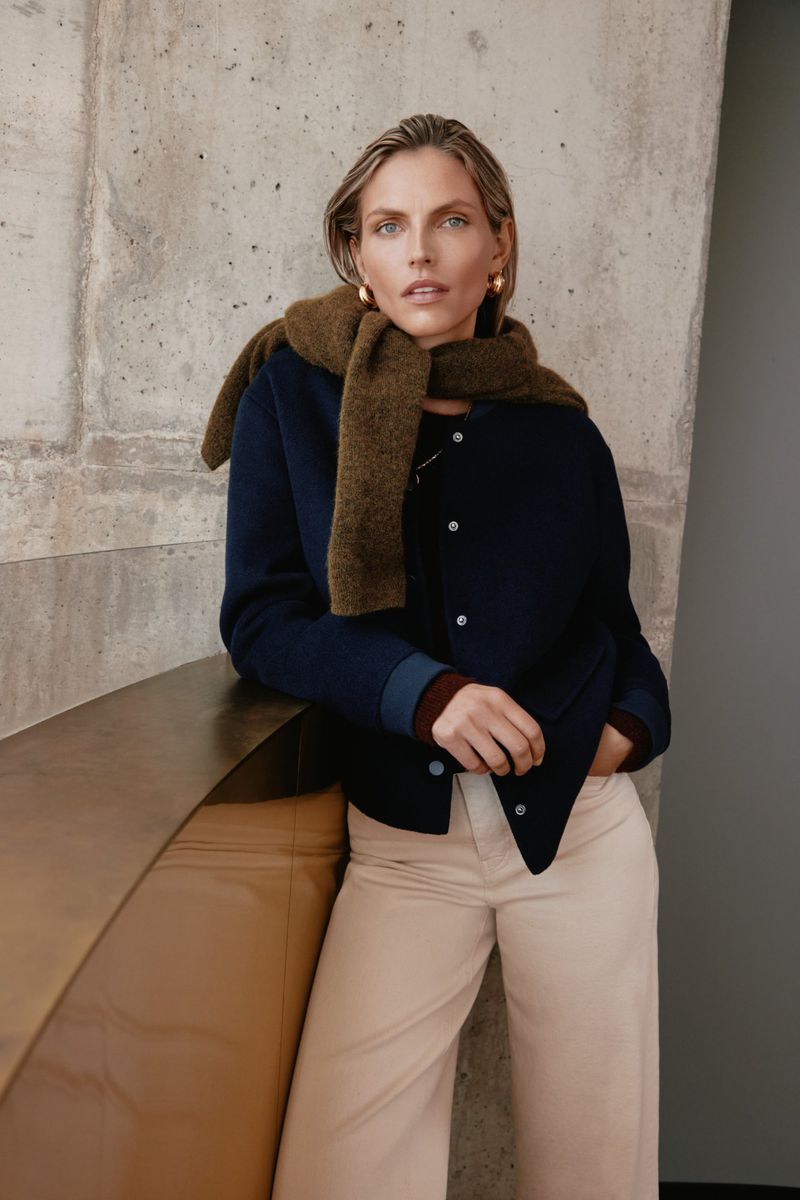 Fashionable woman in navy jacket and tan pants poses against a textured wall with a stylish scarf, showcasing contemporary style.