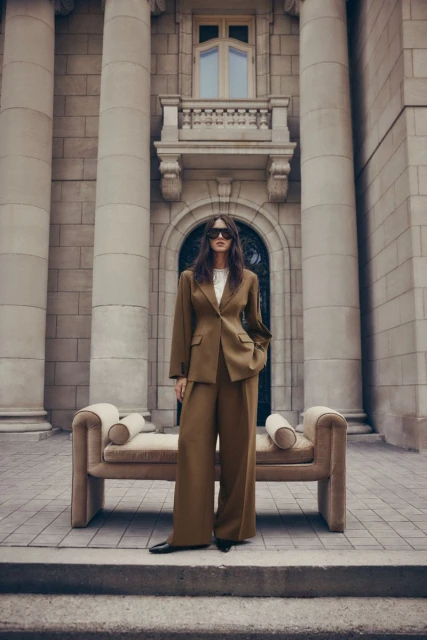 Woman in a stylish brown suit poses on a stone staircase with a long modern sofa, against a building with large columns.