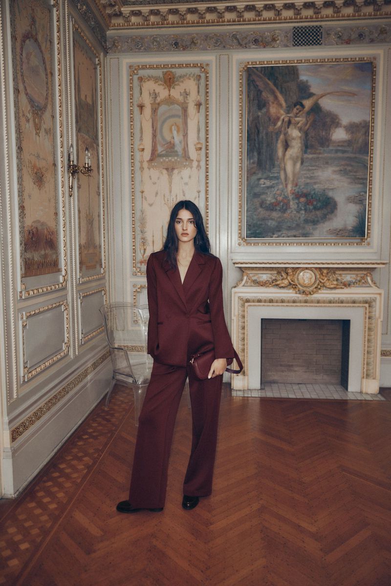 A woman poses in a maroon suit inside a formal room with paneled walls, herringbone floors, and a large painting over a fireplace.