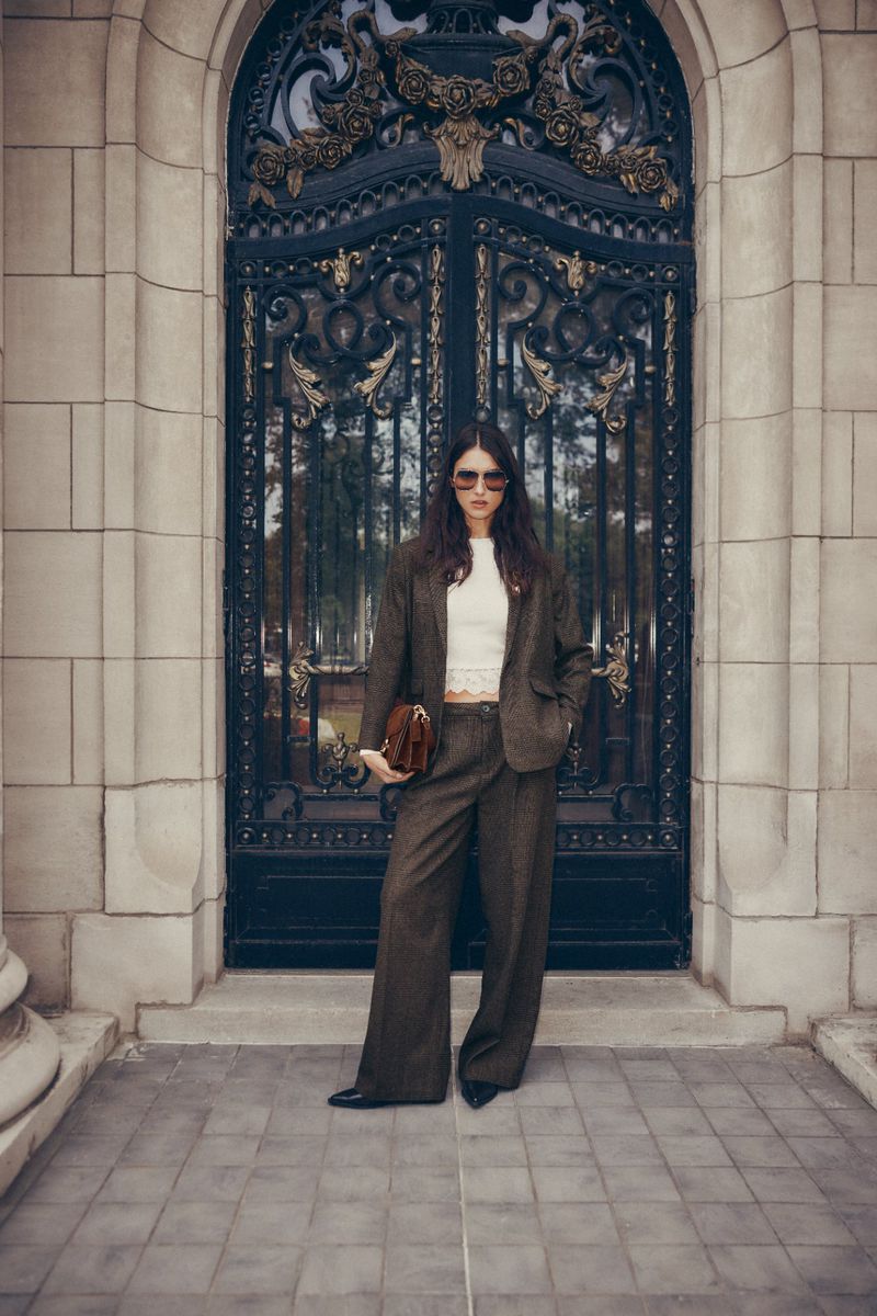 Elegant woman in a brown suit stands before a wrought-iron gate, holding a handbag, showcasing fashion and architecture.