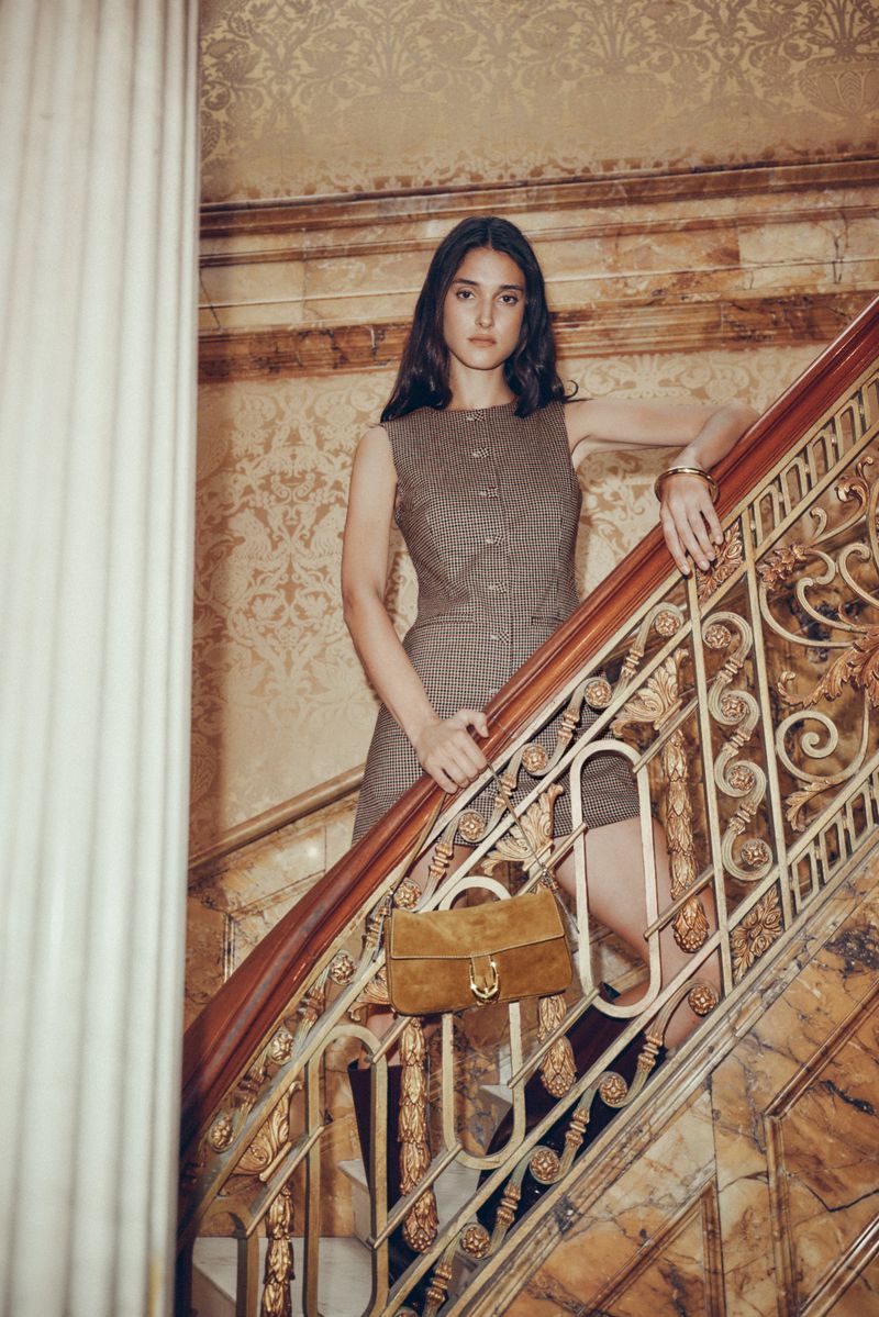 A young woman poses on a marble staircase, wearing a brown sleeveless dress and holding a suede handbag, in an indoor setting.