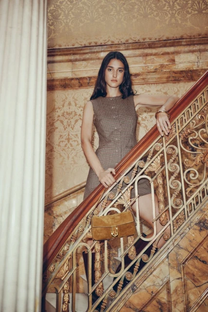 A young woman poses on a marble staircase, wearing a brown sleeveless dress and holding a suede handbag, in an indoor setting.