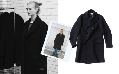Model wearing a dark coat standing against a white brick wall next to a garment rack and a product cutout of a coat