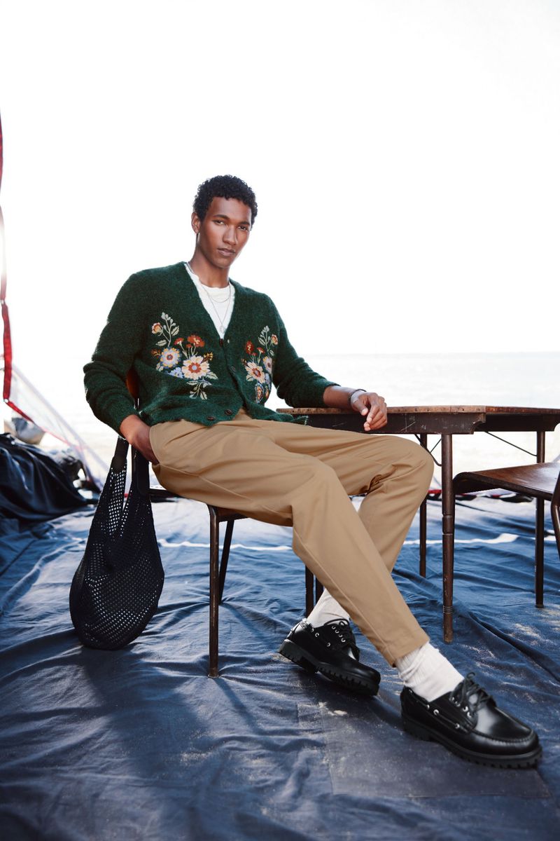A stylish dark-skinned man sits outdoors on a black tarp, wearing a floral-embroidered cardigan, khaki pants, and black loafers.