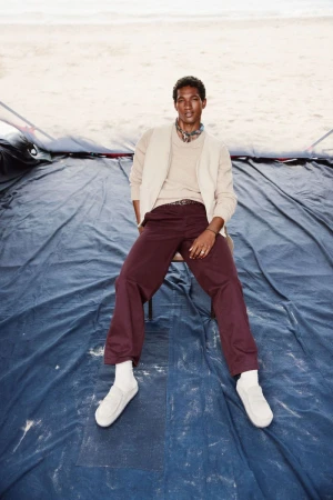 A well-lit, full shot of a man sitting on a chair, wearing a cardigan and trousers on a blue tarp, shot in daylight.