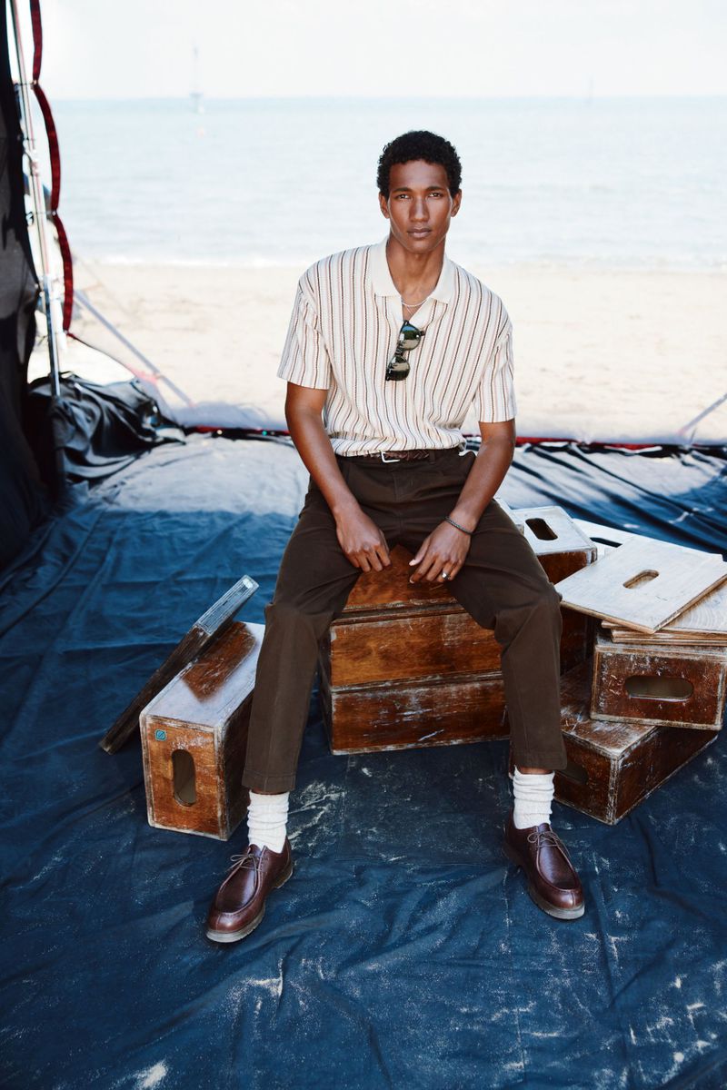 Man in striped shirt sits on wooden boxes near the beach in a fashion shot, wearing brown trousers and shiny shoes in outdoor setting.