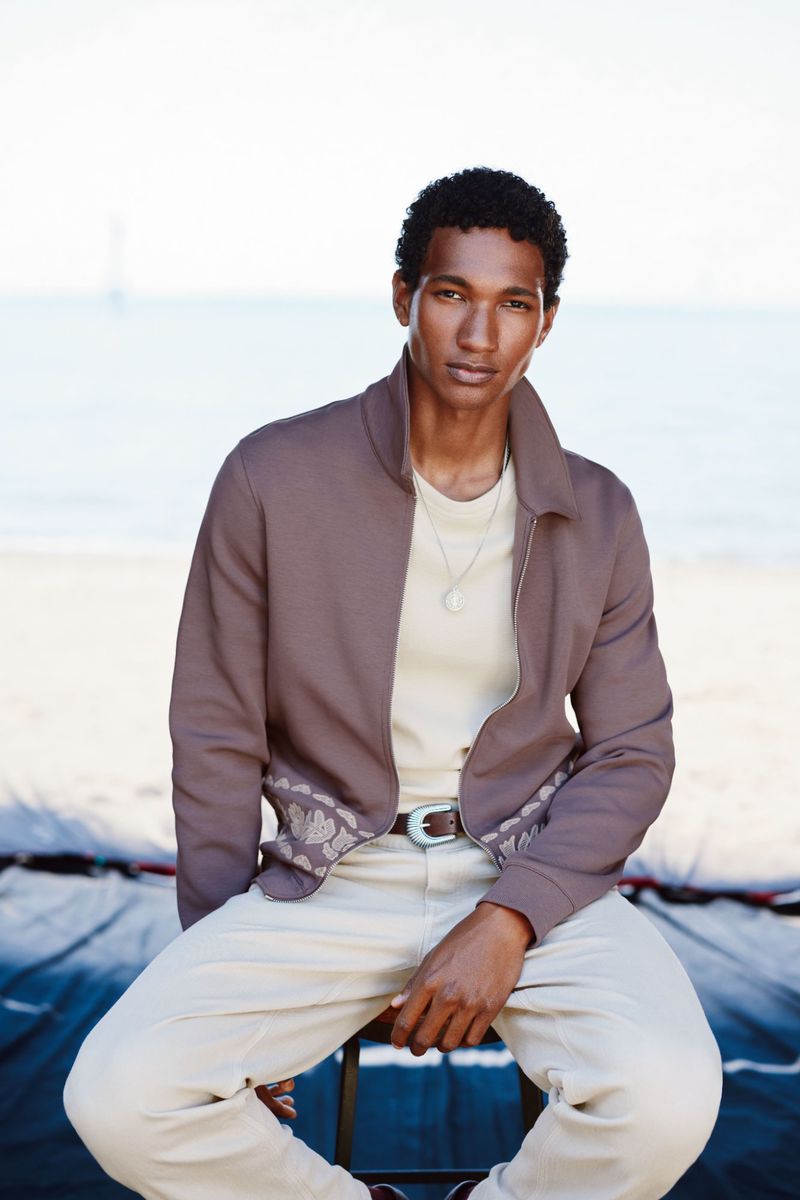 Portrait of a handsome man in a brown jacket and cream pants sitting on a stool in front of a beach on a sunny day.