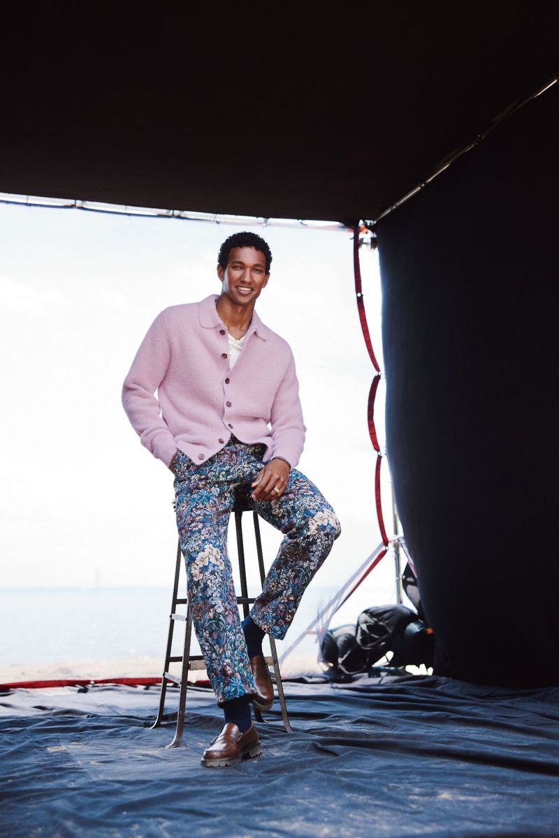 A smiling, dark-haired adult male sits on a stool on a photo set, wearing a pink cardigan and floral pants, with ocean backdrop.