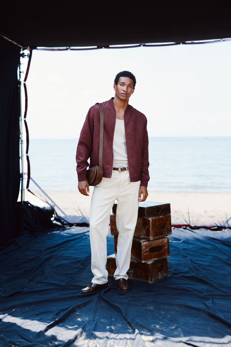 A dark-haired man in a wine-colored jacket and cream pants poses near the ocean on a black platform, standing in front of stacked chests.