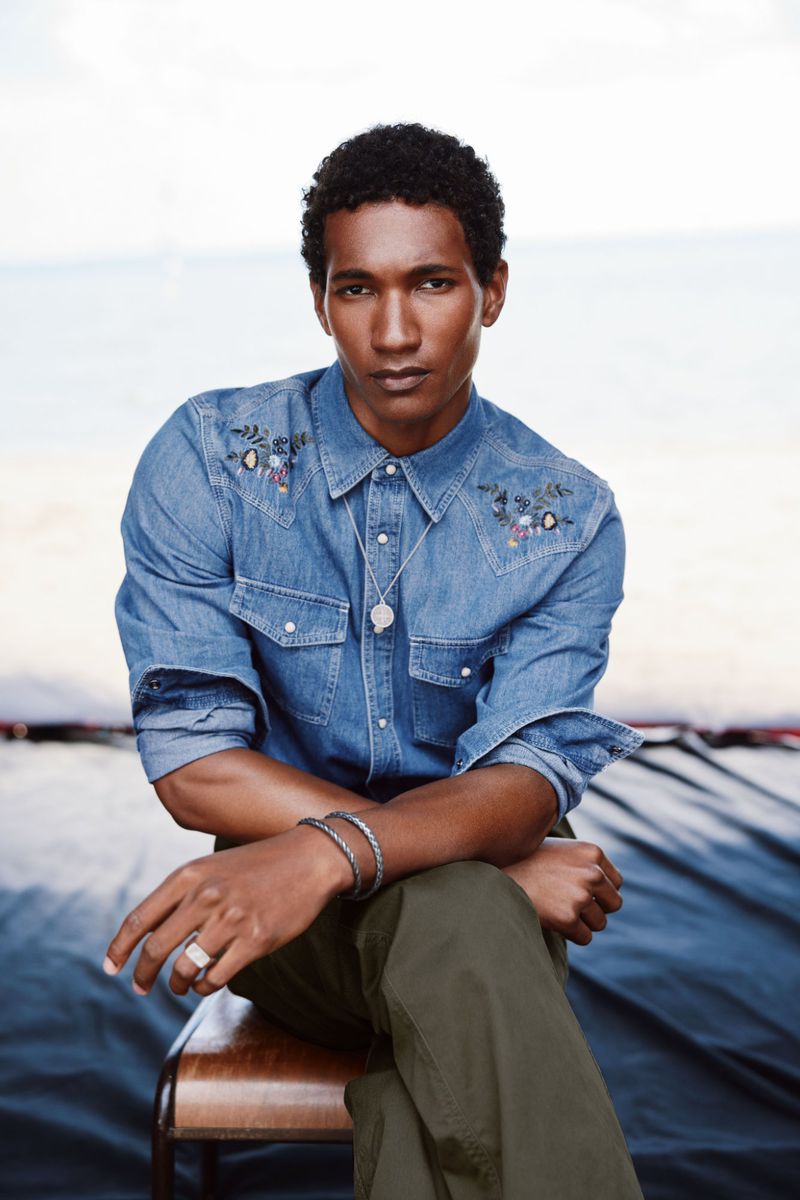 A man is photographed from the waist up, sitting on a chair, wearing a blue denim shirt with floral embroidery and olive-green pants.