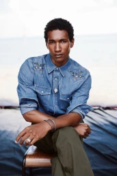 Model Joshua Seth wearing an embroidered denim shirt and olive trousers seated on a chair outdoors.