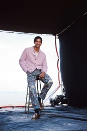 Model Joshua Seth sitting on a stool wearing a light pink cardigan and blue floral trousers on an outdoor photoshoot set.