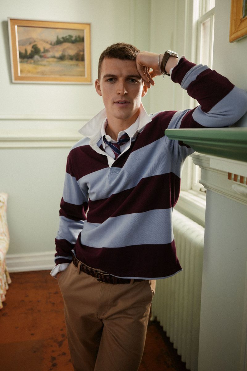 A young man with short brown hair is casually leaning against a white mantle wearing a maroon and light blue striped polo shirt indoors.