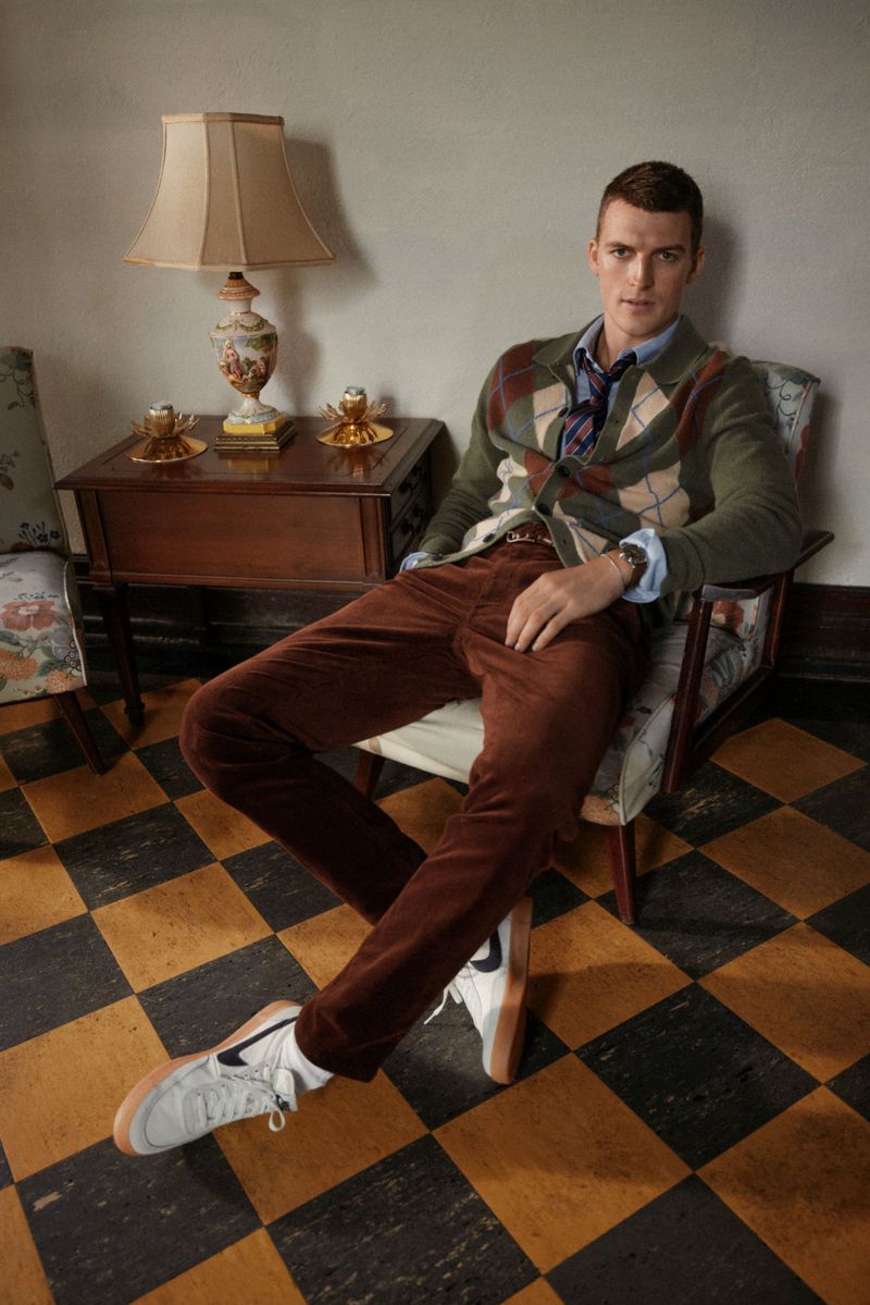 A fair-skinned man in a sweater and brown pants sits in a chair indoors near a table and lamp on a checkered floor.