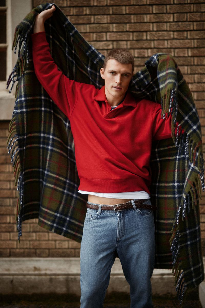 Portrait of a young adult male wearing a red sweater and jeans, holding a large plaid blanket in front of a brick wall.