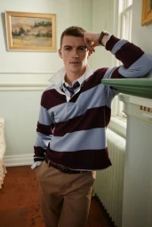 Model leaning against a mantelpiece wearing a burgundy and blue striped rugby shirt and khaki trousers.